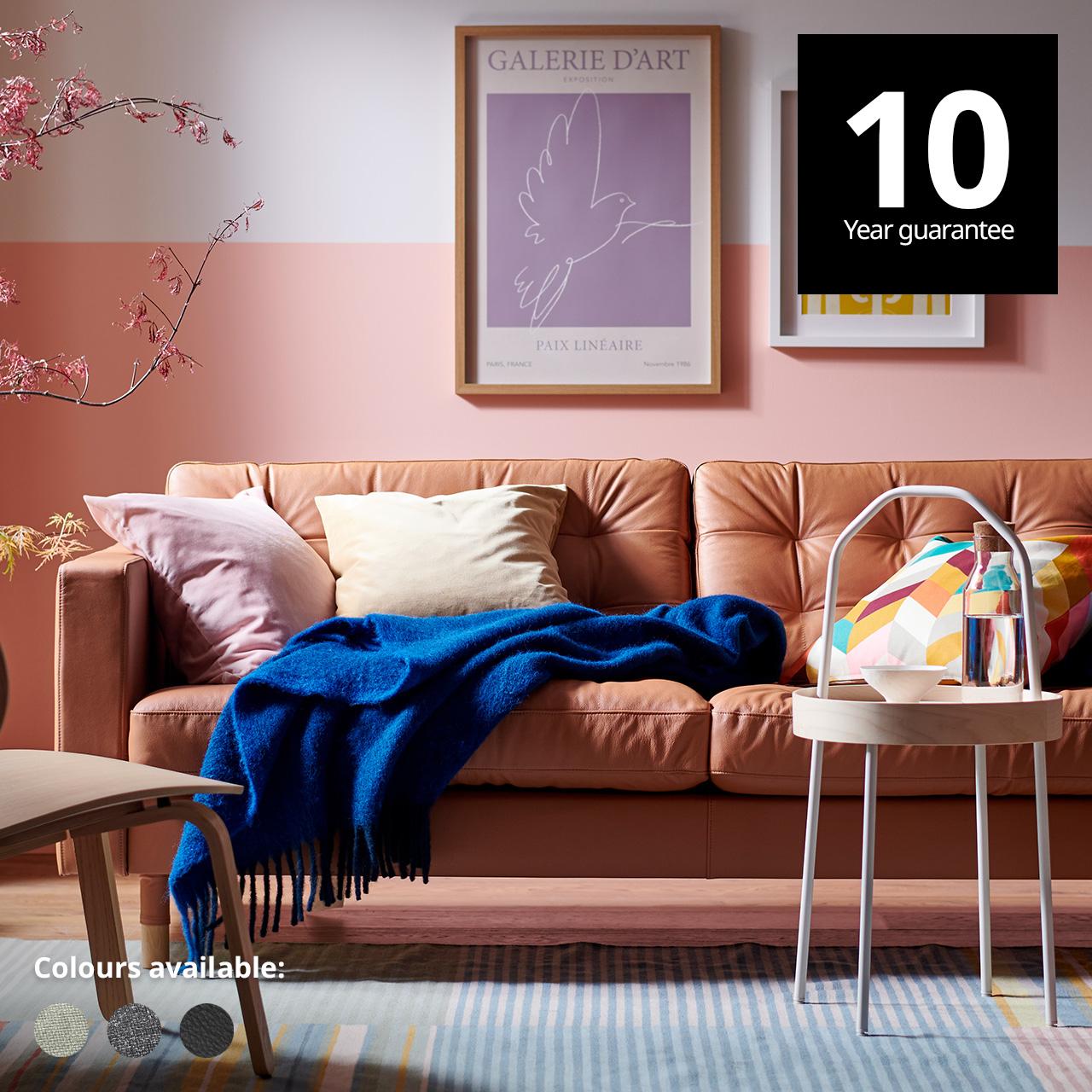 A LANDSKRONA sofa, with a blue KLIPPOXEL throw and extra cushions on it, and a BURVIK side table stand on a BUDDINGE rug. A 10-year guarantee tag is placed over the image at the top right corner. White text overlay image that reads "Colours available:" with the colour swatch of Gunnared light green, Gunnared dark grey and Bomstad black.