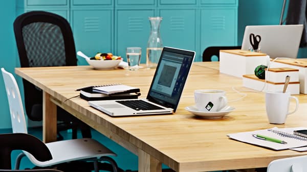 A laptop is opened on a NORDEN table with notebooks and a cup of drinks.
