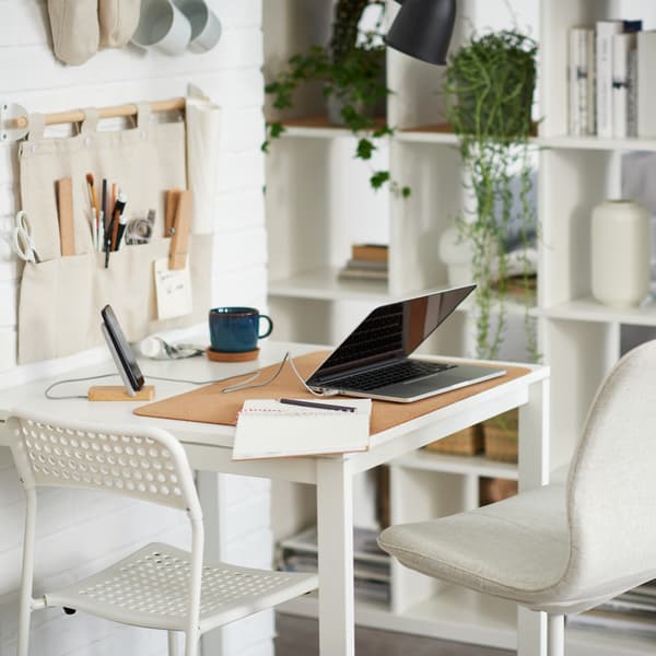 A laptop on a white table with one white and one beige chair, beside a white shelving unit with books, plants and storage.