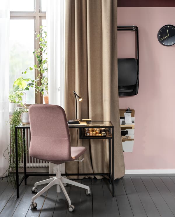 A laptop table in black-brown/glass and a light brown-pink conference chair stand by a window with beige and white curtains.