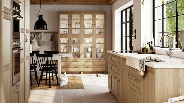 A large, bright kitchen with oak VEDHAMN fronts, including glass door cabinets filled with glassware and dinnerware.