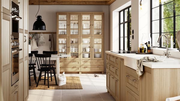 A large, bright kitchen with oak VEDHAMN fronts, including glass door cabinets filled with glassware and dinnerware.