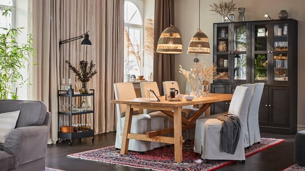 A large dining room with a wooden table, chairs with beige chair covers, dark brown cabinets, beige curtains, a trolley.
