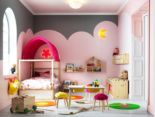 A large kids' bedroom furnished with a pine bed with posts and a pink canopy. Shown together with a pine table that has two storage boxes under the table top and a storage bench with plastic boxes in white and pink.