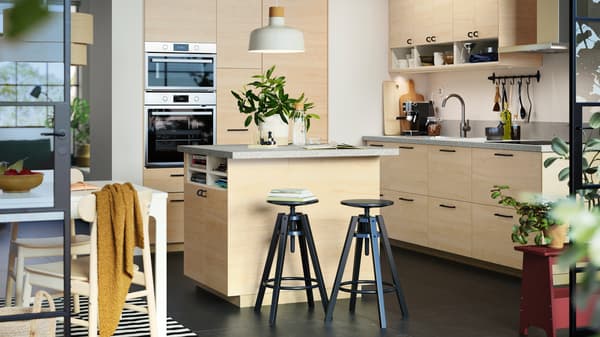 A large kitchen island with bright FRÖJERED fronts in light bamboo has a white pendant lamp above it and two black stools.