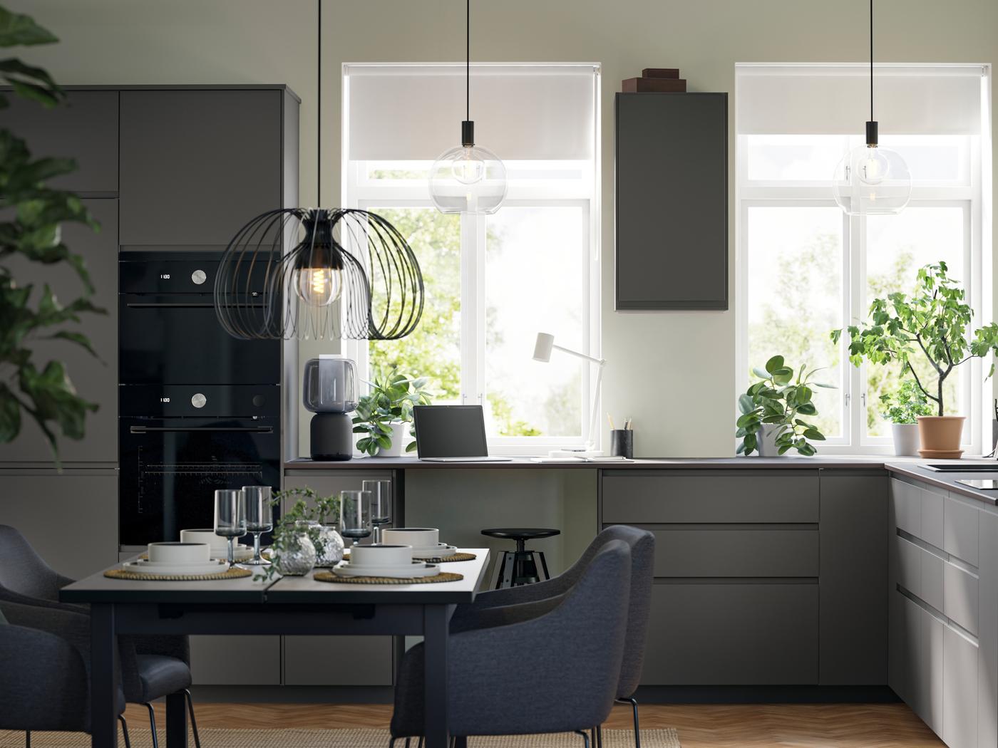 A large kitchen with dark grey VOXTORP fronts and black appliances has two large windows and a dining area with four chairs.