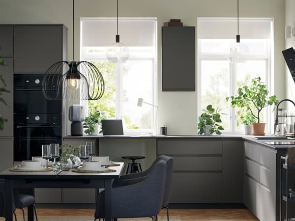 A large kitchen with dark grey VOXTORP fronts and black appliances has two large windows and a dining area with four chairs.