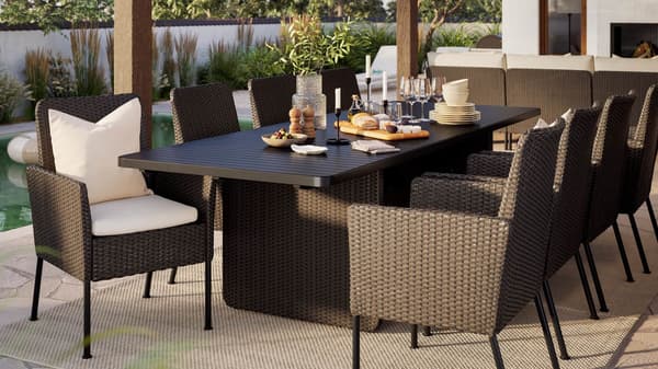 A large outdoor space with dark gray VITTSKÄR table and eight rattan dining chairs under a roof. Sun is shining.