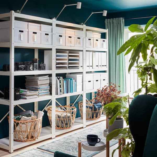 A large white BROR shelving unit with storage boxes, cameras, magazines, books, magazine files and three baskets in rattan.