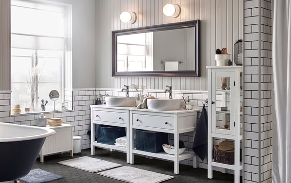 A large white tiled bathroom, with wood panelled walls, with two wash-stands and wash-basins side by side, below a large mirror.