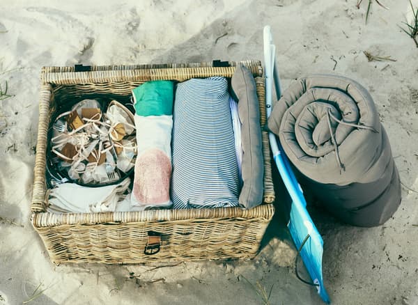 A large wicker basket full of party essentials, a cushion and foladable chair sit in the sand.
