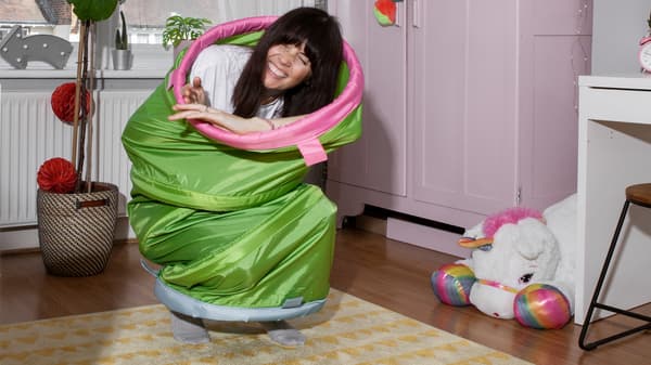 A laughing woman has crept into a BUSA play tunnel and half sits, half stands on the carpet in a children’s room.
