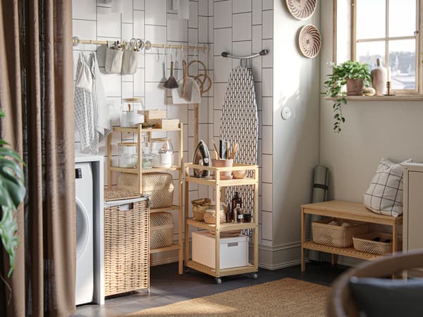 A laundry nook with storage boxes and laundry accessories on open shelving and a RÅGRUND trolley, against a white-tiled wall.