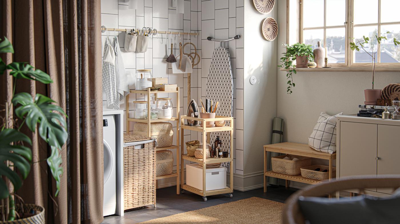 A laundry nook with storage boxes and laundry accessories on open shelving and a RÅGRUND trolley, against a white-tiled wall.