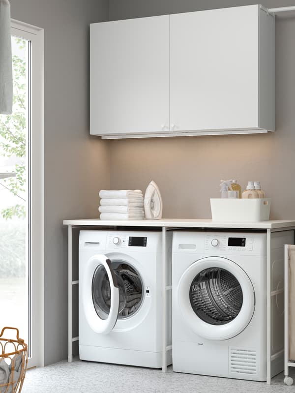 A laundry with a washing machine and a tumble dryer next to an ENHET laundry bag with castors and some wall cabinets above.