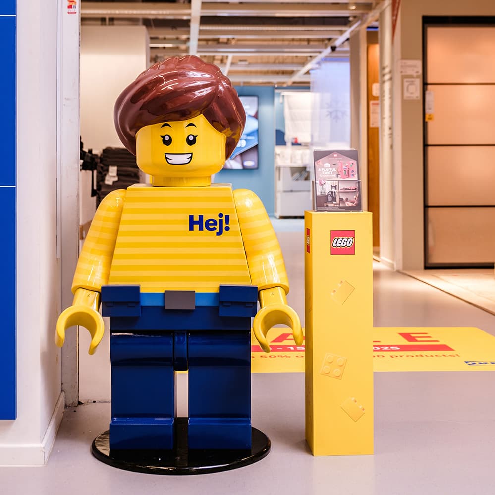 A life-size LEGO display figure dressed in the iconic IKEA uniform: yellow striped shirt and blue pants.