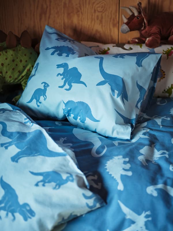 A light and dark blue dinosaur quilt and pillow sit on a child’s bed, while two JÄTTELIK dinosaurs peek out.