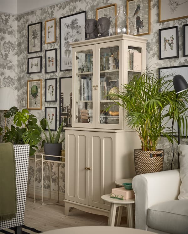 A light beige cabinet with both panel and glass doors stands in front of a wall with leaf-patterned wallpaper.