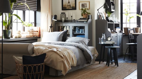A light beige sofa-bed made as a bed with grey bedding. Behind is a grey glass-door cabinet and a jute rug lies on the floor.