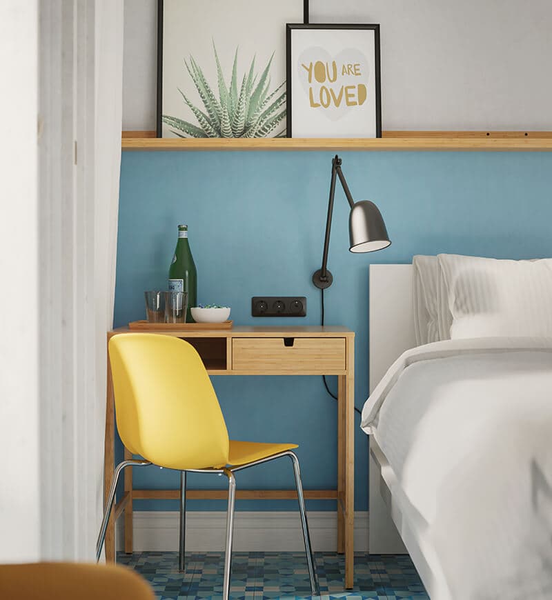 A light blue room featuring a white bed with a small wooden desk and yellow chair beside it.