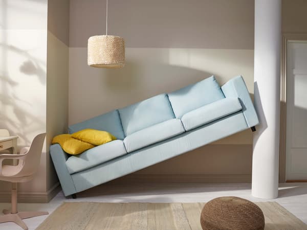 A light blue sofa with yellow cushions on top, tilted between two walls in a beige room, a beige rug on the floor.