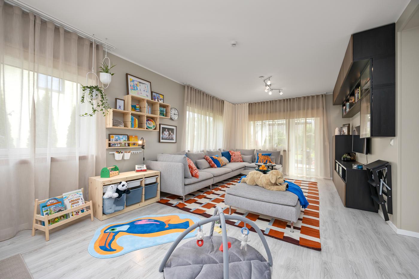 A light-filled living room with a large window and sheer curtains. A grey sofa with colorful cushions sits on a patterned rug. The walls are decorated with wall shelves and framed pictures.