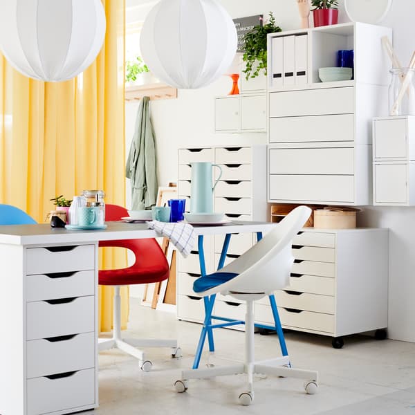 A light gray/white/blue table with drawer unit, two swivel chairs in white/blue/red, white drawer units and yellow curtains.