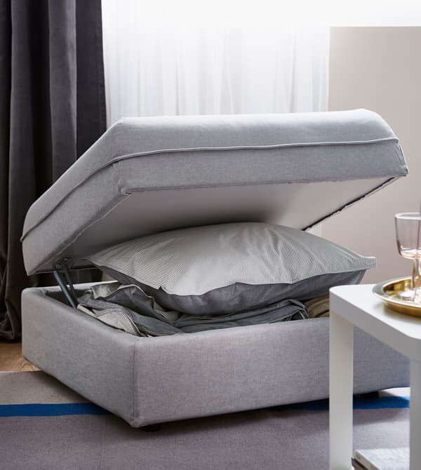 A light grey IKEA VALLENTUNA modular sofa unit, with lid opened to reveal a storage compartment for pillows and bedding.