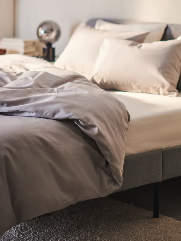 A light grey SLATTUM upholstered bed frame with an ÅKREHAMN foam mattress and NATTSVÄRMARE bed linen stands against a wall.