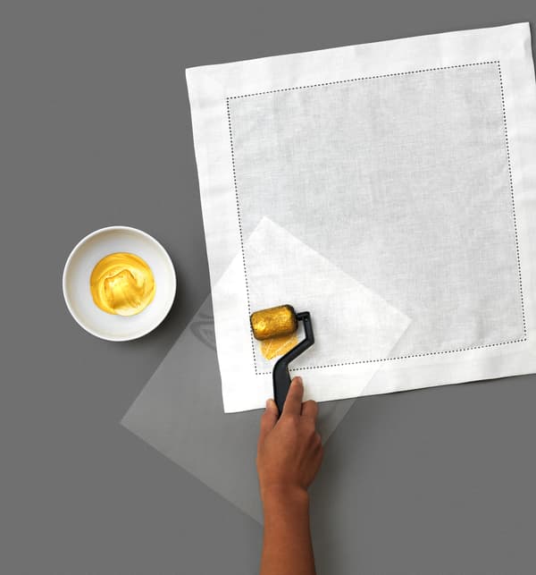 A little DIY goes a long way to make custom, decorative napkins using white GULLMAJ cloth napkins from IKEA. The third step is to place the cut-out plastic stencil on the napkin. Then use a roller to apply fabric paint onto the napkin.