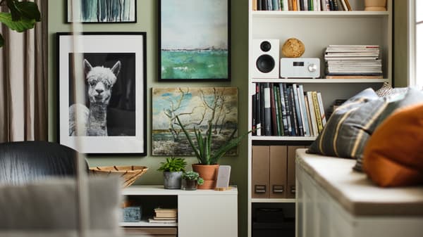 A living area with a white BILLY bookcase holding books and a speaker, against a wall with framed prints and photographs.