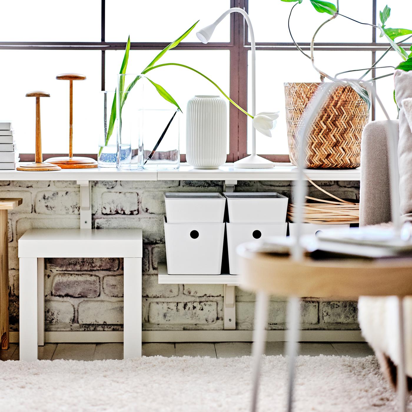 A living area with white BERGSHULT/RAMSHULT wall shelves with white KUGGIS boxes with lids and a white LACK side table.