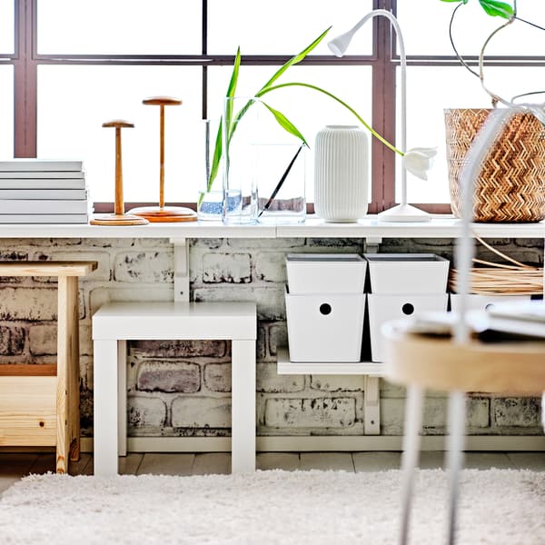 A living area with white BERGSHULT/RAMSHULT wall shelves with white KUGGIS boxes with lids and a white LACK side table.