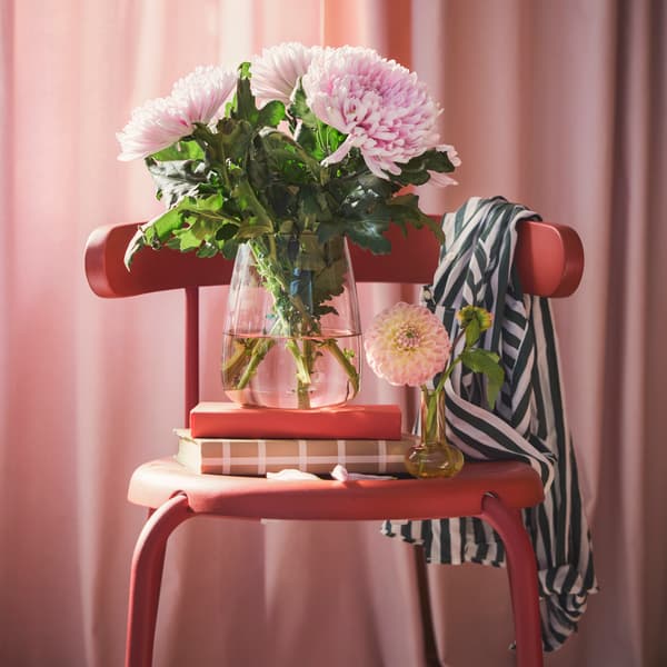 A living room features a light pink BERÄKNA vase made of glass filled with large pink flowers on a red chair beside books.