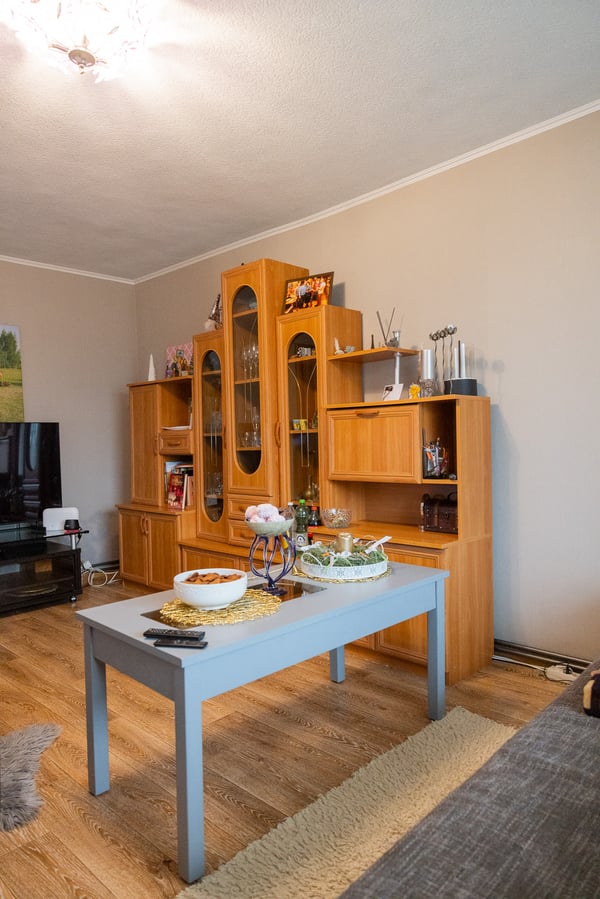 A living room featuring a wooden wall unit, a television, and a small table in the centre. Open shelving and various household items create a busy appearance.