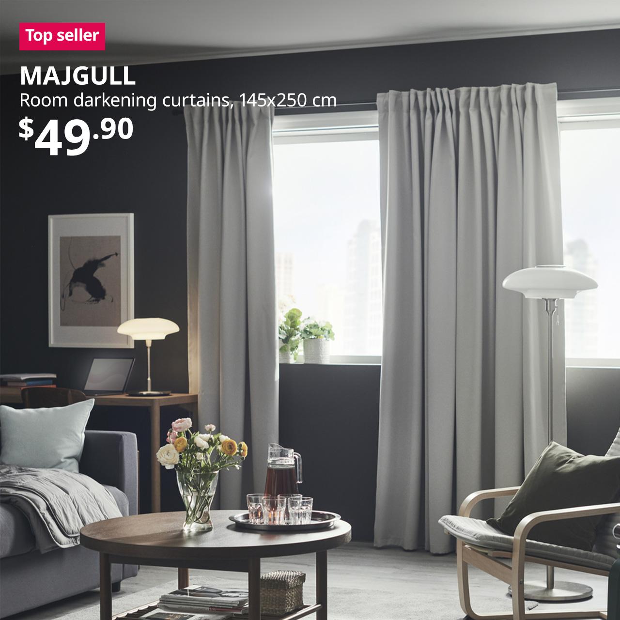 A living room setting using a pair of light grey MAJGULL room-darkening curtains. A reddish-pink tag with white text that reads "Top Seller", followed by a white text overlay image that reads "MAJGULL, Room darkening curtains, 145x250 cm, $49.90".