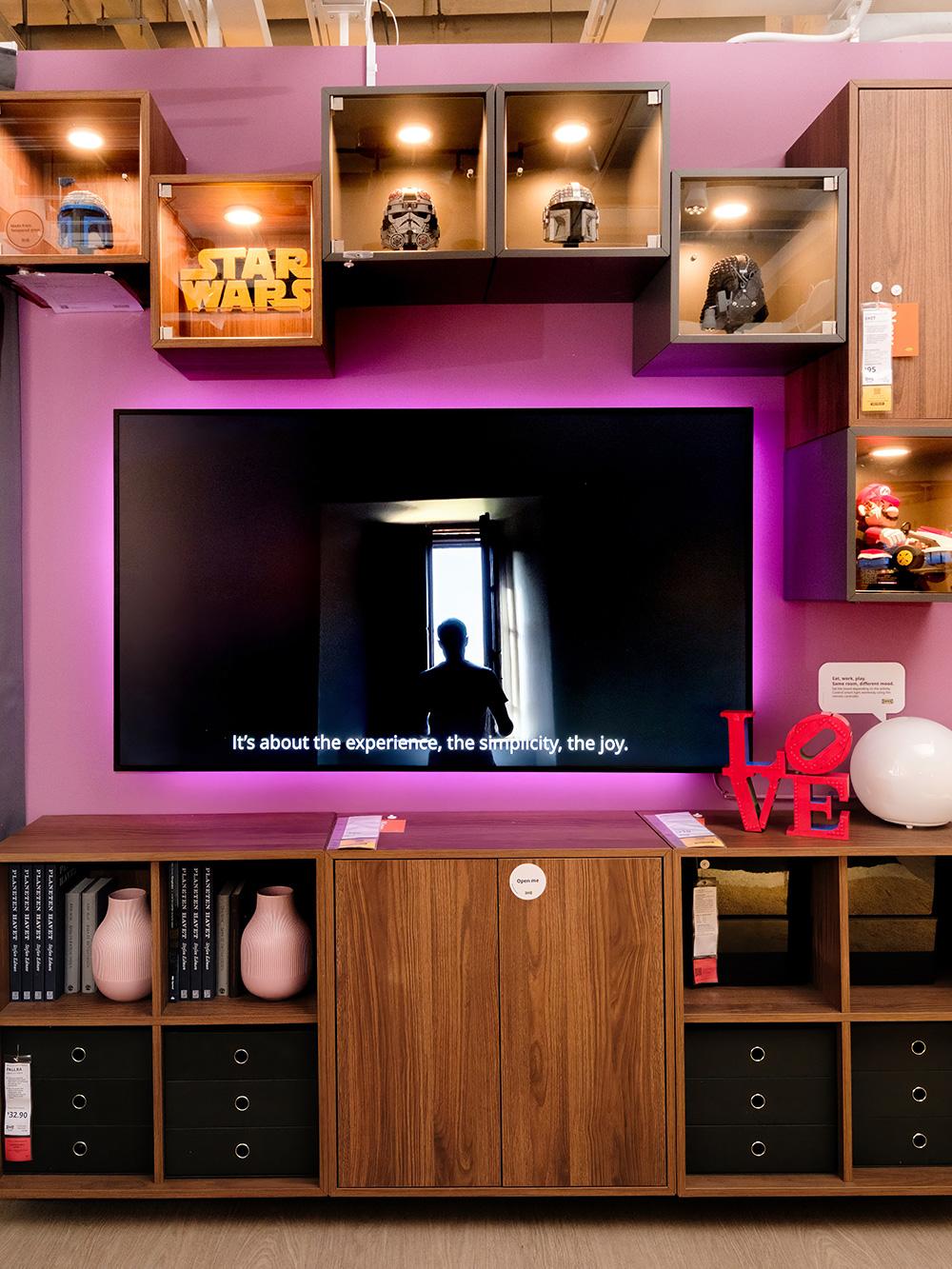 A living room setting with brown/walnut effect EKET storage series is shown in this display alongide with a smart LED backlight on a 65" TV. Items in the showroom are as followed: A EKET cabinet with 4 compartments on both sides of a EKET cabinet with 2 doors and 1 shelf. Five EKET wall cabinet with glass door is mounted above the TV, two in brown walnut effect and three in dark grey with LEGOs displayed with MITTLED LED spotlight highlighting the display.