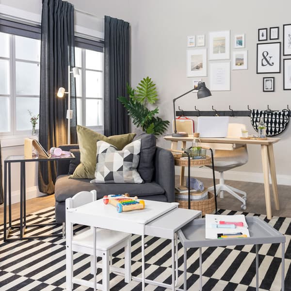 A living room shows nesting tables being used for kids' play. A sofa with a laptop stand is behind the nesting tables and a workspace is set up towards the back of the living room.