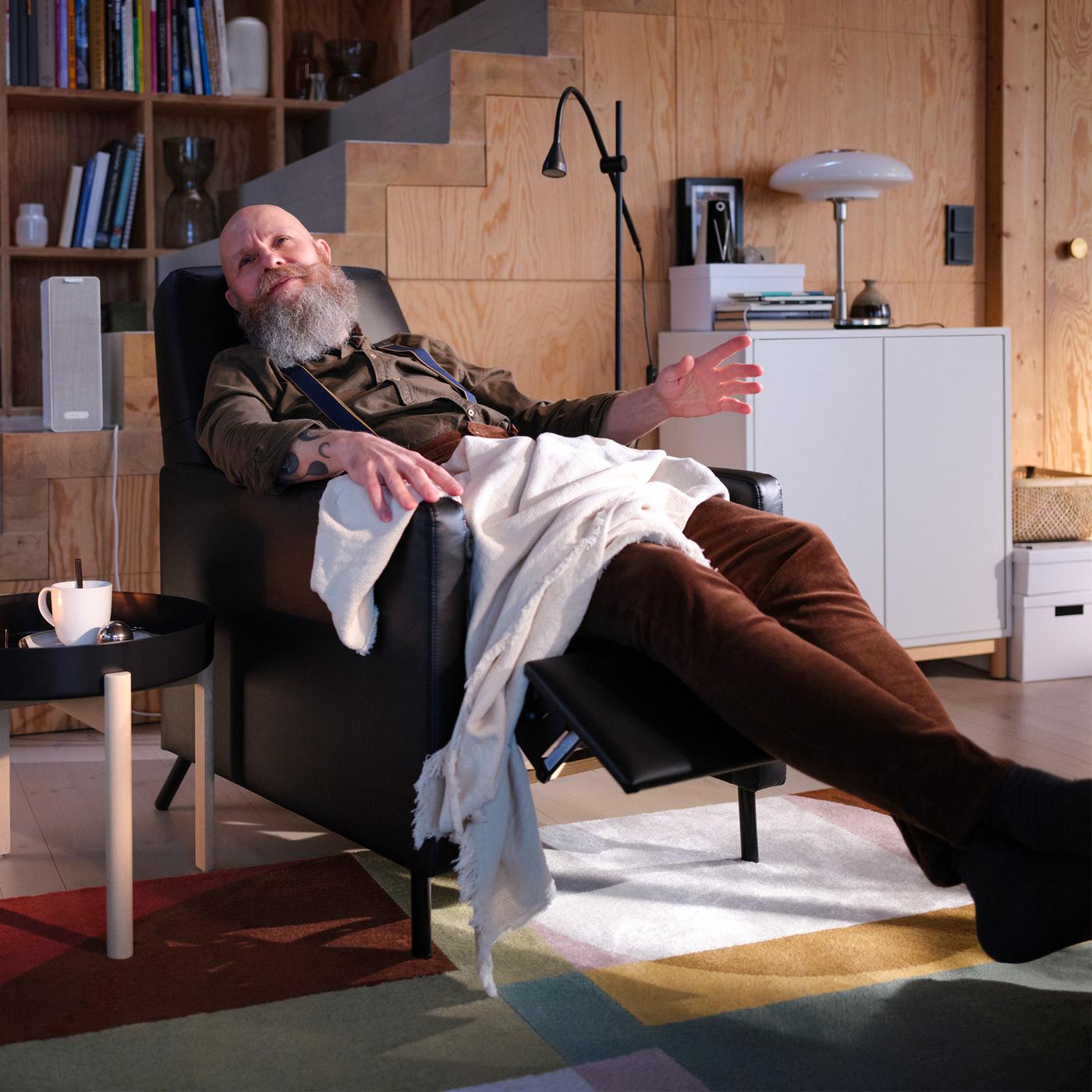 A living room with a man sitting in a black GISTAD recliner listening to music beside a coffee table with a cup of tea.
