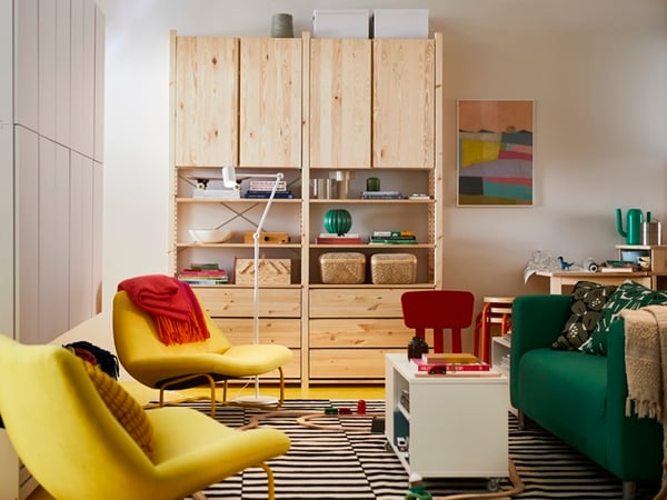 A living room with a natural wood storage unit against the wall, a green sofa, a yellow armchair draped with a red blanket, a striped rug, a white coffee table, and colourful artwork on the wall, creating a warm and inviting atmosphere.