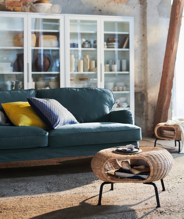 A living room with a rattan and metal GAMLEHULT footstool, a dark green STOCKSUND sofa and glass-fronted cabinets.