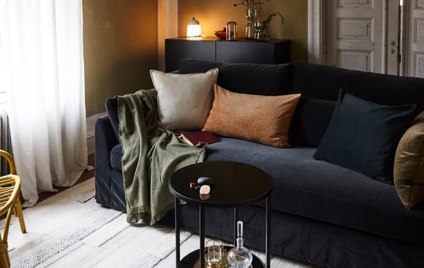 A living room with a sofa, cushions, throw, rug, a small table, a sideboard with a lamp, vases, and a rattan armchair.