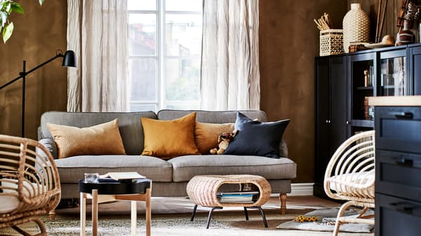 A living room with a sofa, two jute rugs, two rattan armchairs, beige linen curtains and cabinets.