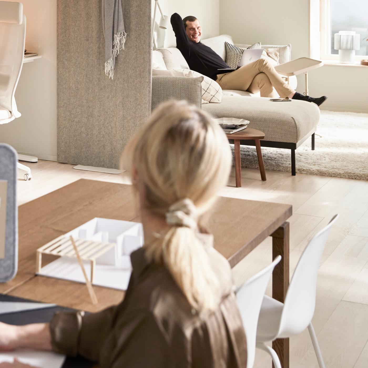 A living room with a woman using ÖVNING desk divider to study, a corner office with BEKANT desk, and a man is sat on a sofa.