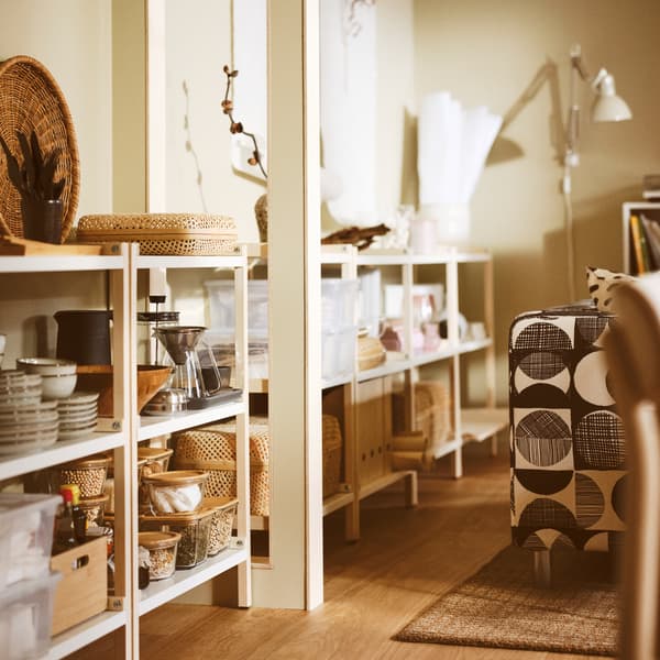 A living room with EKENABBEN open shelving units containing GLADELIG bowls, a HÖGMODIG coffee maker, and boxes with lids.