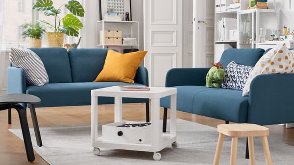 A living room with two 2-seat sofas, a white side table on castors, a children's stool, cushions, potted plants.