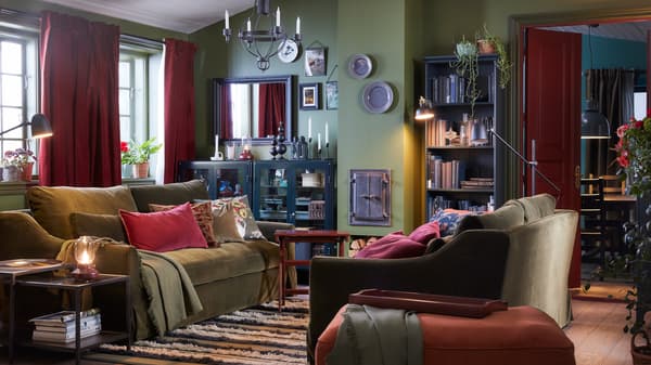 A living room with two olive-green sofas, a light red footstool, black chandelier, red-brown curtains and a striped rug.