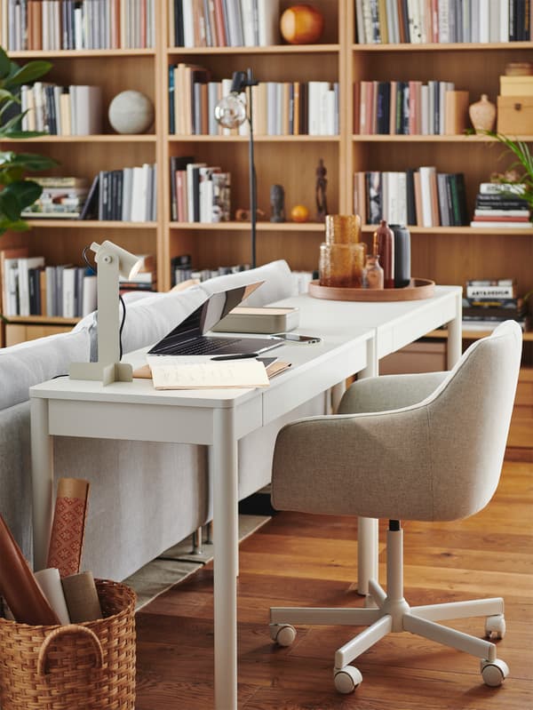 A living space with a small workstation placed behind a sofa, consisting of a white TONSTAD desk and a FLOTTILJ lamp.