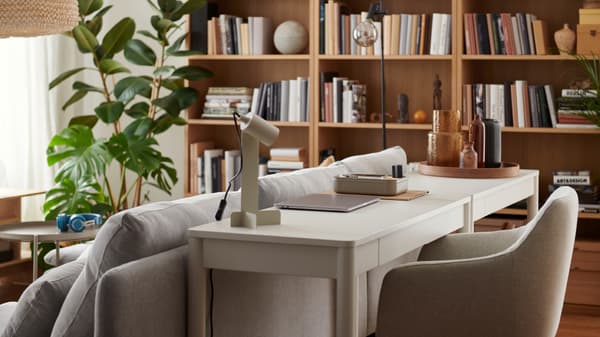A living space with a small workstation placed behind a sofa, consisting of a white TONSTAD desk and a FLOTTILJ lamp.