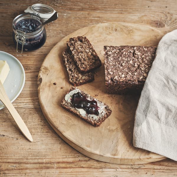 A loaf of KNÅDA multigrain bread with cut slices on a wooden plate placed on a wooden table with butter and jam.